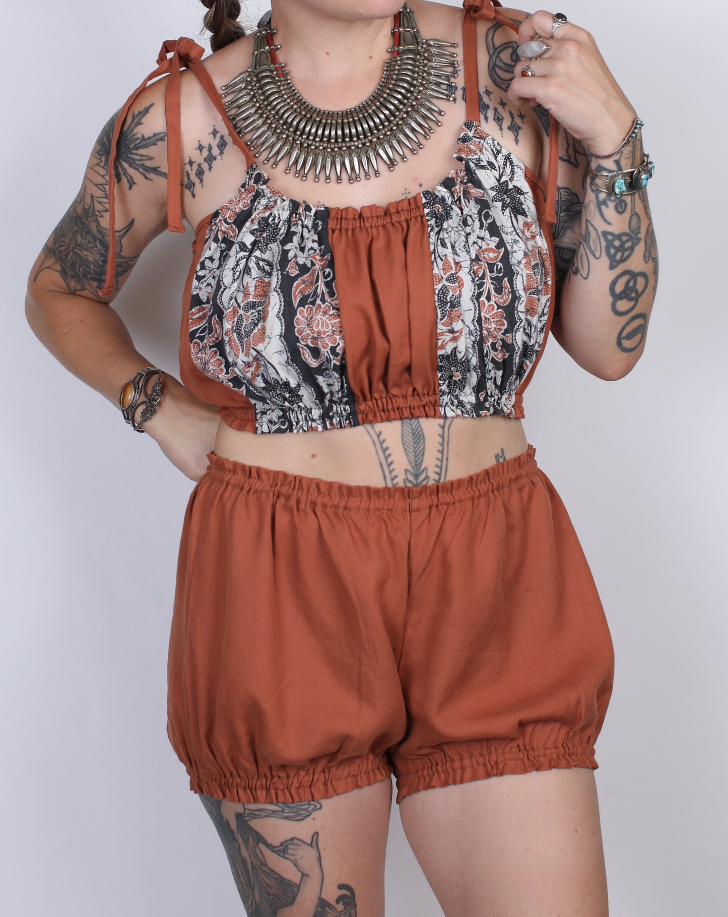 A woman modeling a two-piece outfit consisting of a rust-orange and black-and-white floral print crop top with tie straps, and matching rust-orange high-waisted balloon shorts. She is accessorized with a large silver statement necklace and bracelets, set against a plain light gray background.